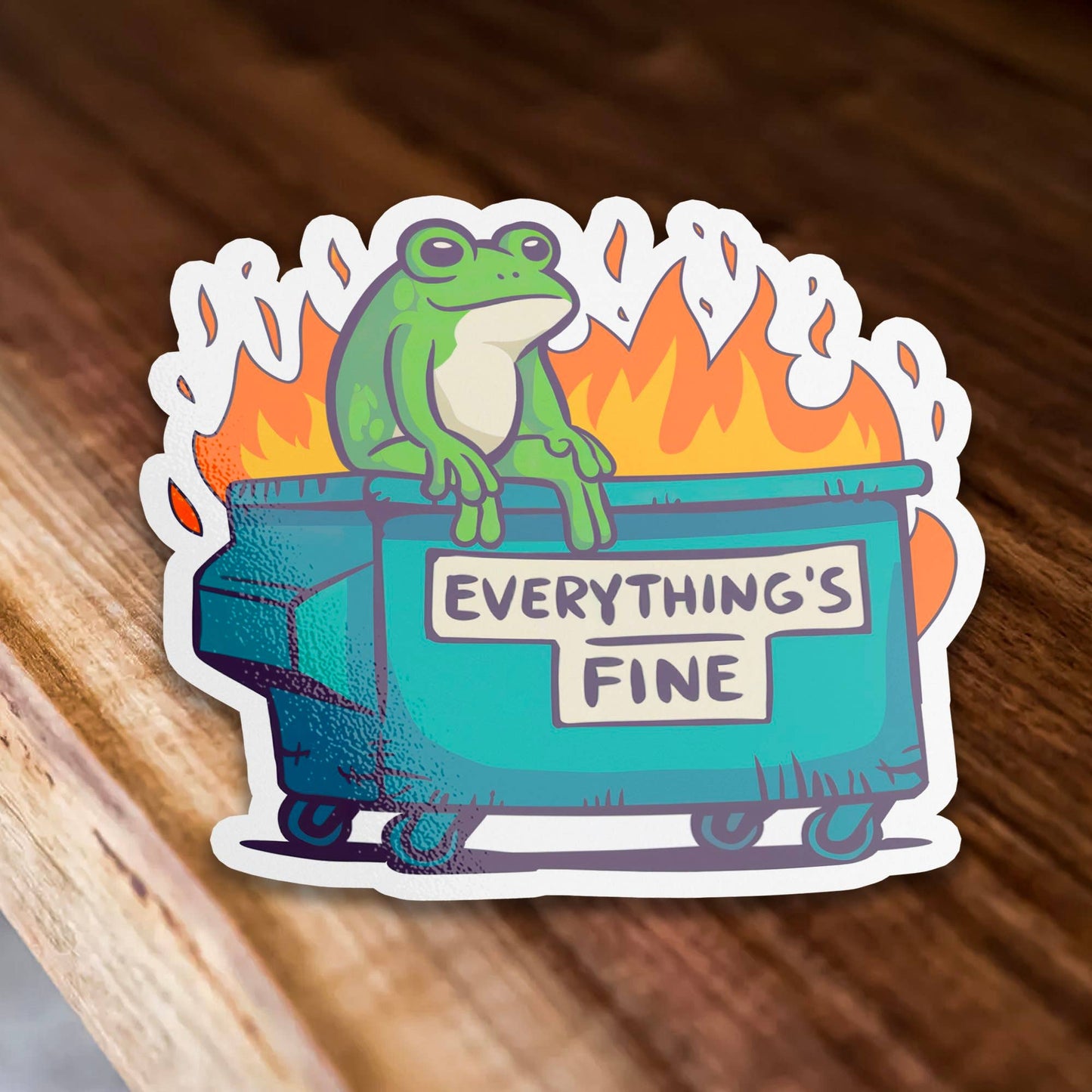 Sticker with a frog on a burning dumpster labeled 'Everything's Fine' on a wooden surface