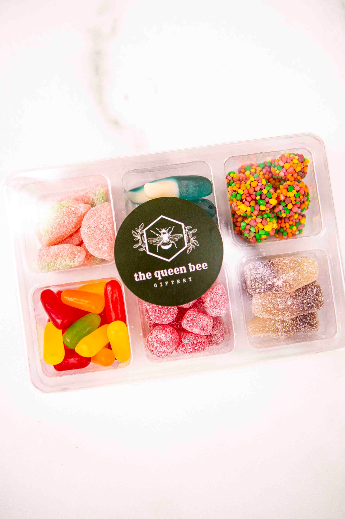 Assorted candies in a divided container with 'the queen bee' label on a white background