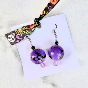Purple Halloween earrings with a witch on a broom on a decorative card with a lanyard.