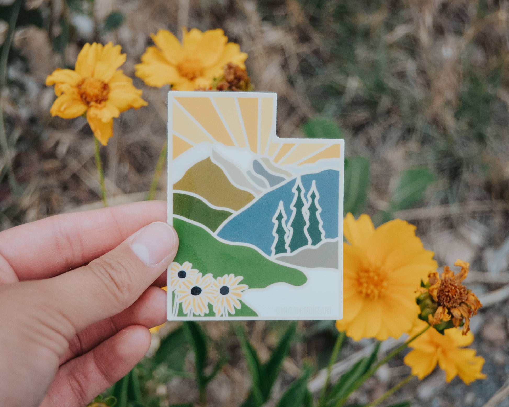 Hand holding a small card with a mountain and flower design, surrounded by yellow flowers.