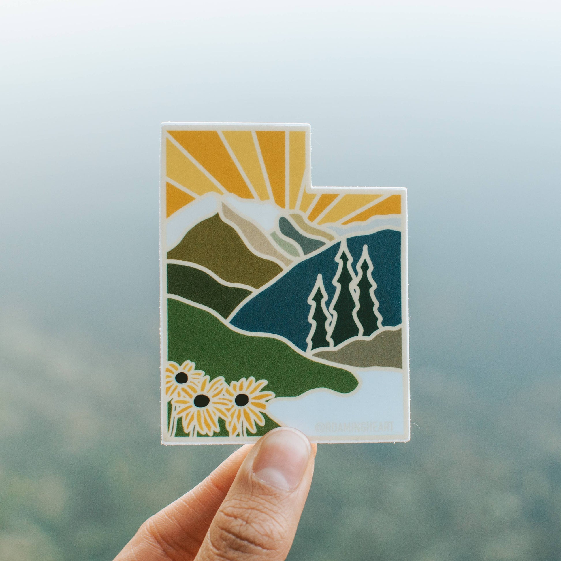 Hand holding a landscape-themed sticker with mountains, trees, and flowers against a blurred natural background.