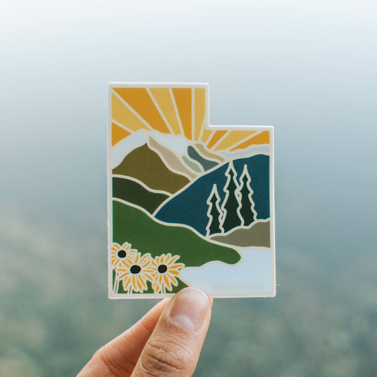 Hand holding a landscape-themed sticker with mountains, trees, and flowers against a blurred natural background.