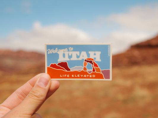 Hand holding a 'Welcome to Utah' sticker with desert landscape in the background