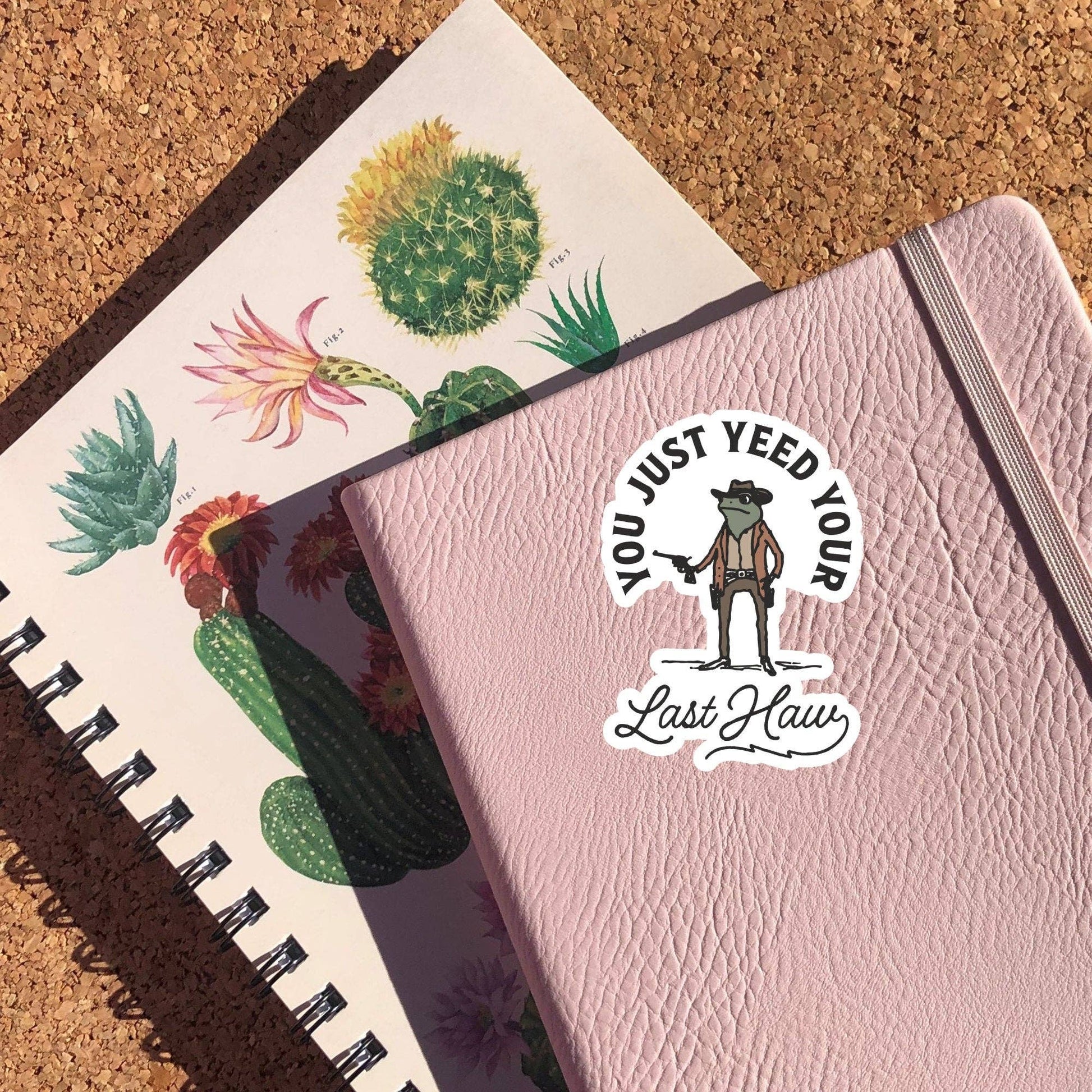 Pink notebook displaying a sticker with a cowboy frog that reads "you just need your last haw"