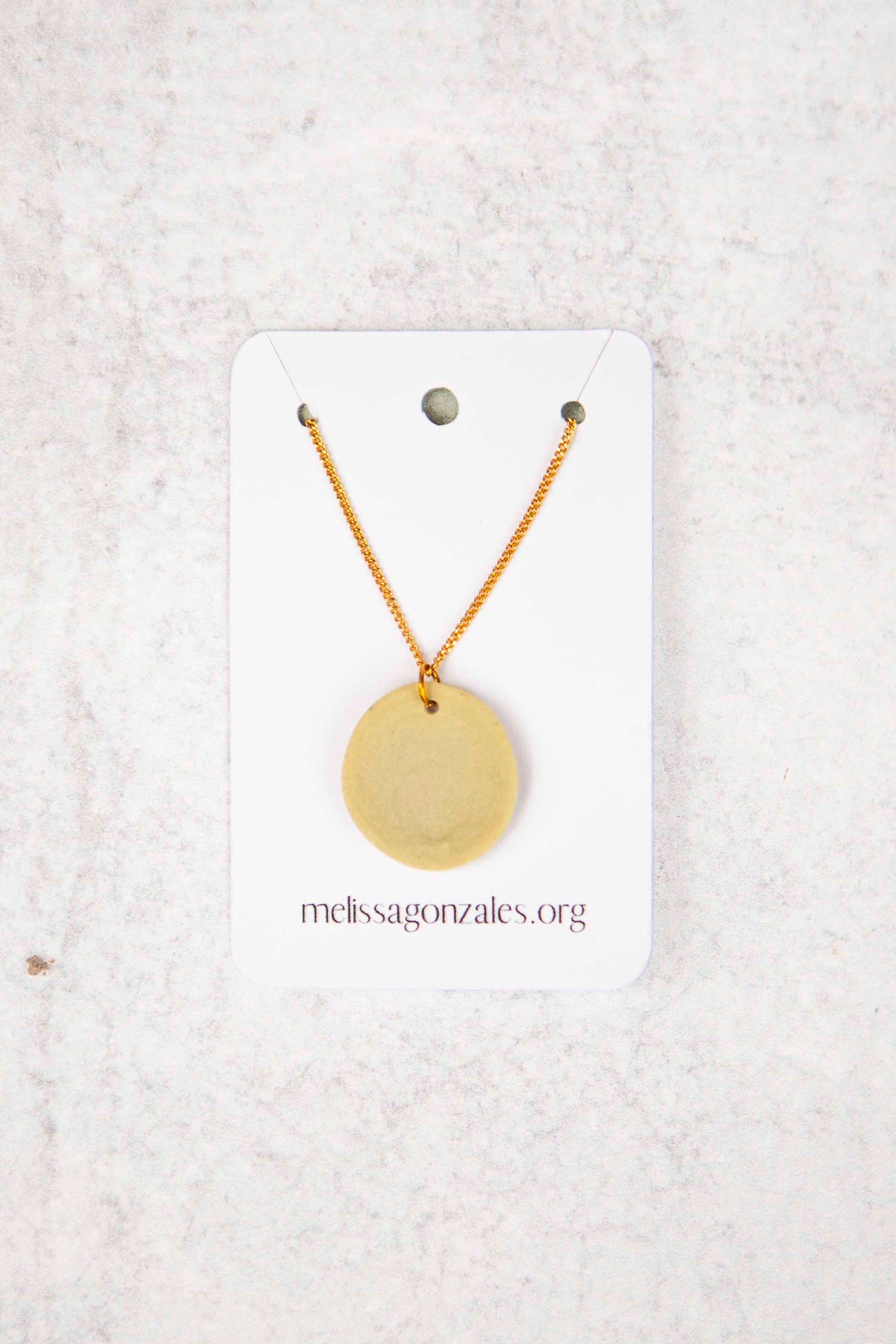 Gold necklace with a round, beige clay pendant on a white card with 'melissagonzales.org' text on a textured white background
