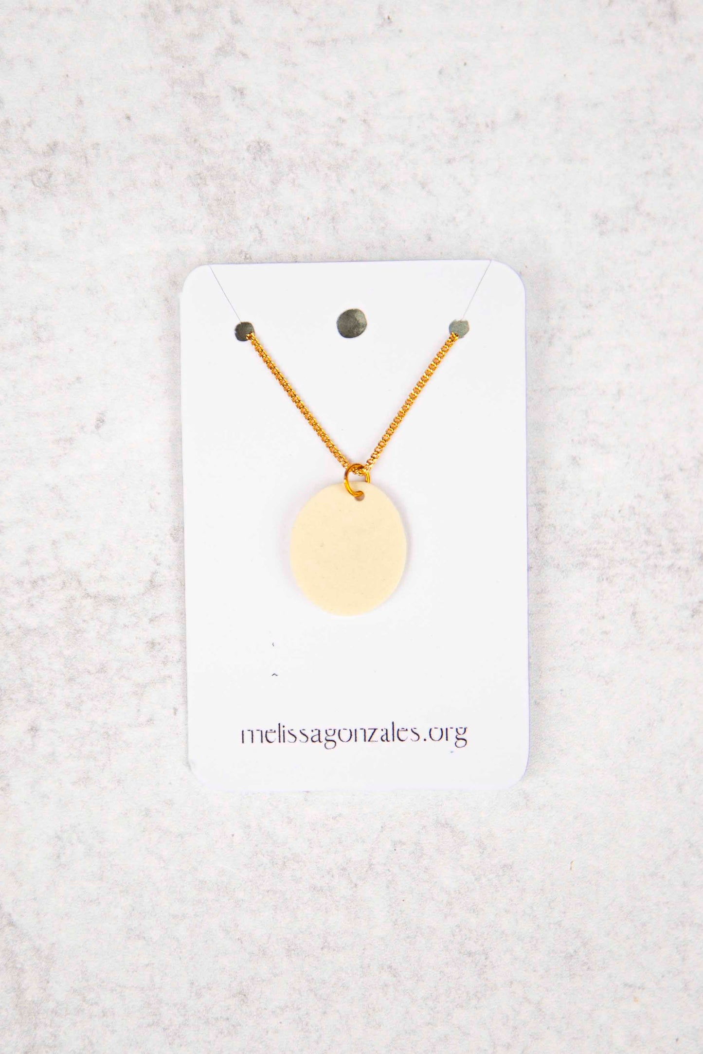 Necklace with a round, cream clay pendant on a white card with 'melissagonzales.org' printed on it, against a light gray background.