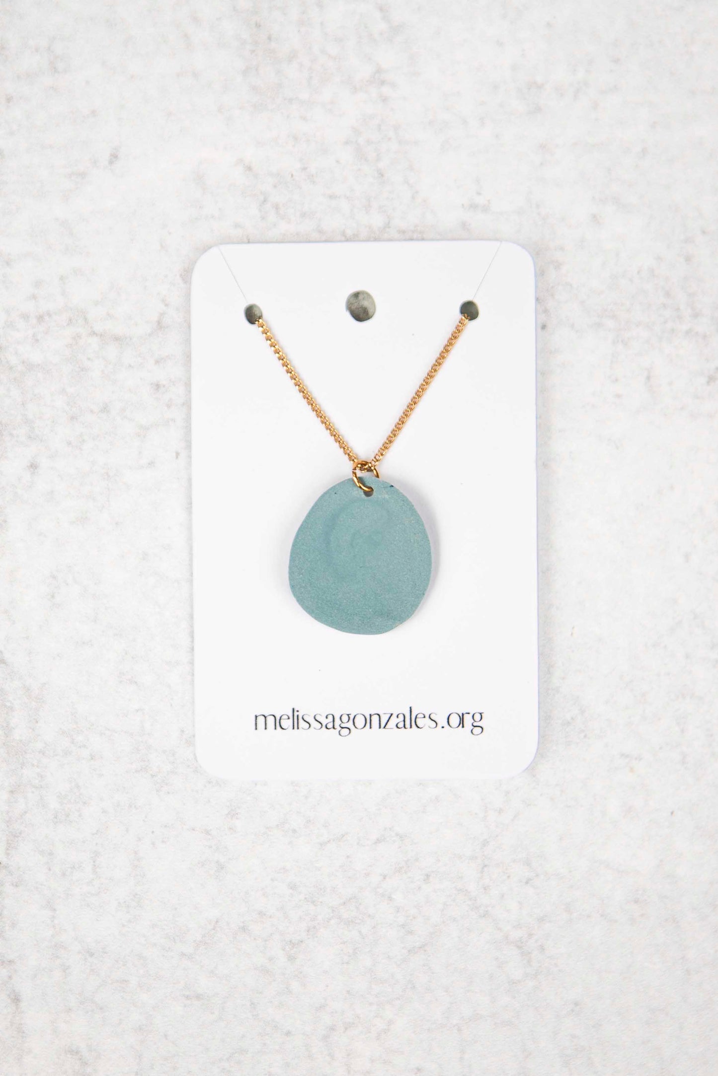 Necklace with a dusty blue, round clay pendant on a white card with 'melissagonzales.org' text, against a textured white background.