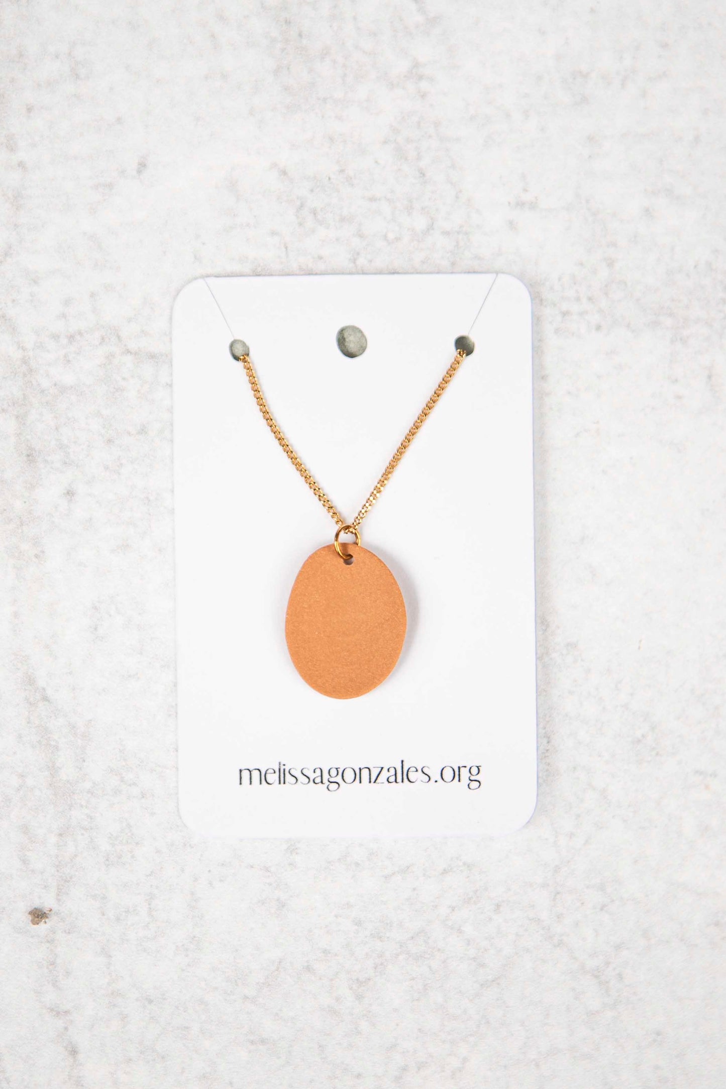 Necklace with a dusty orange round pendant on a white card with 'melissagonzales.org' text, against a light gray textured background.