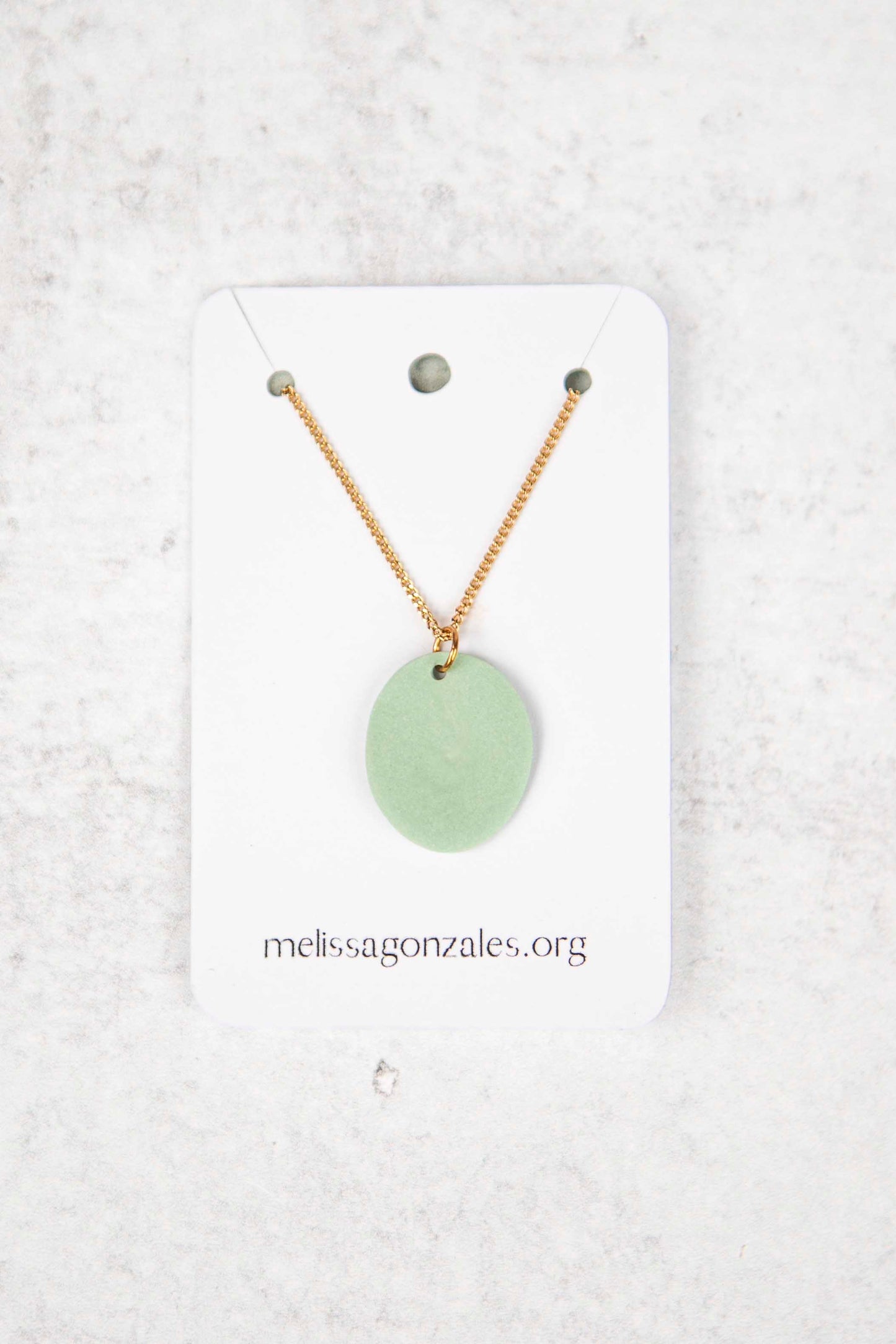 Necklace with a mint colored pendant on a white card with 'melissagonzales.org' printed on it, against a light gray textured background.