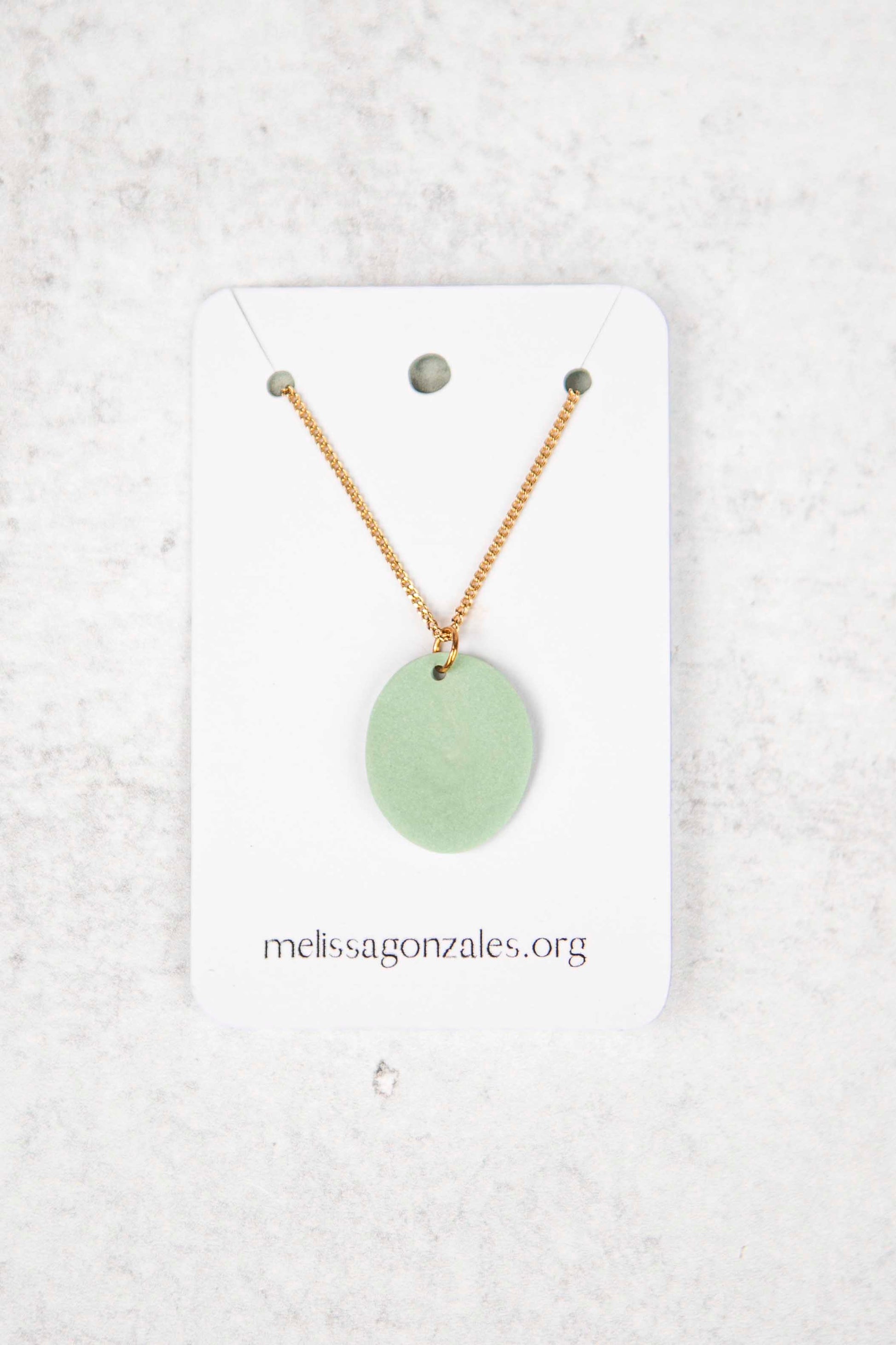 Necklace with a mint colored pendant on a white card with 'melissagonzales.org' printed on it, against a light gray textured background.