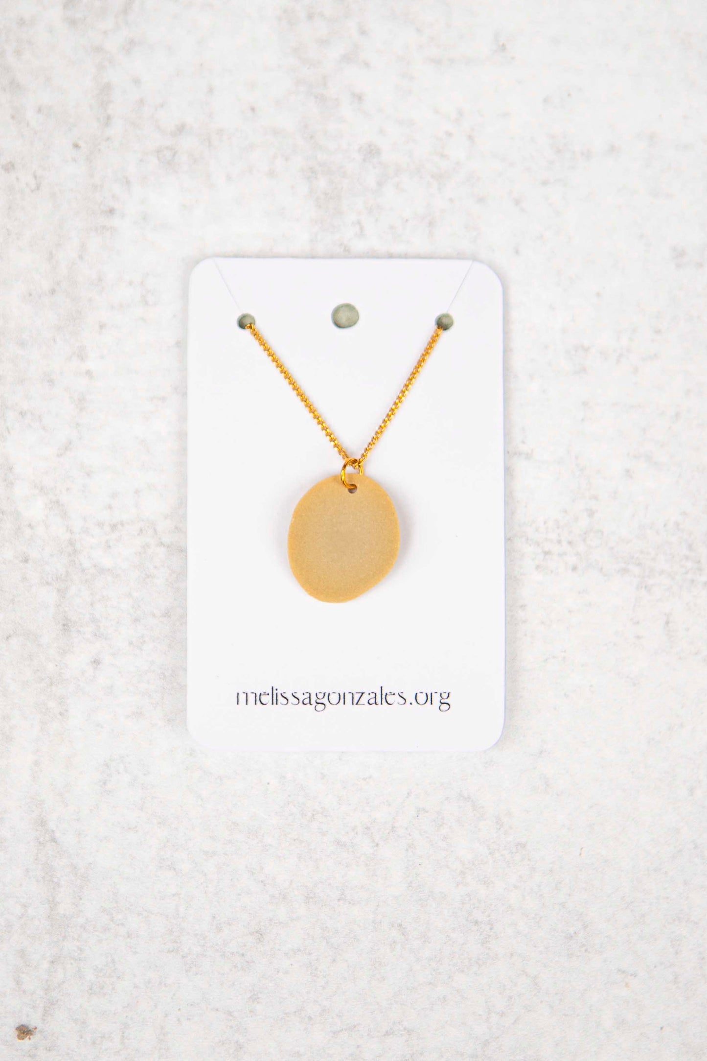 Gold necklace with a round, sand colored clay pendant on a white card with 'melissagonzales.org' text, against a textured white background.
