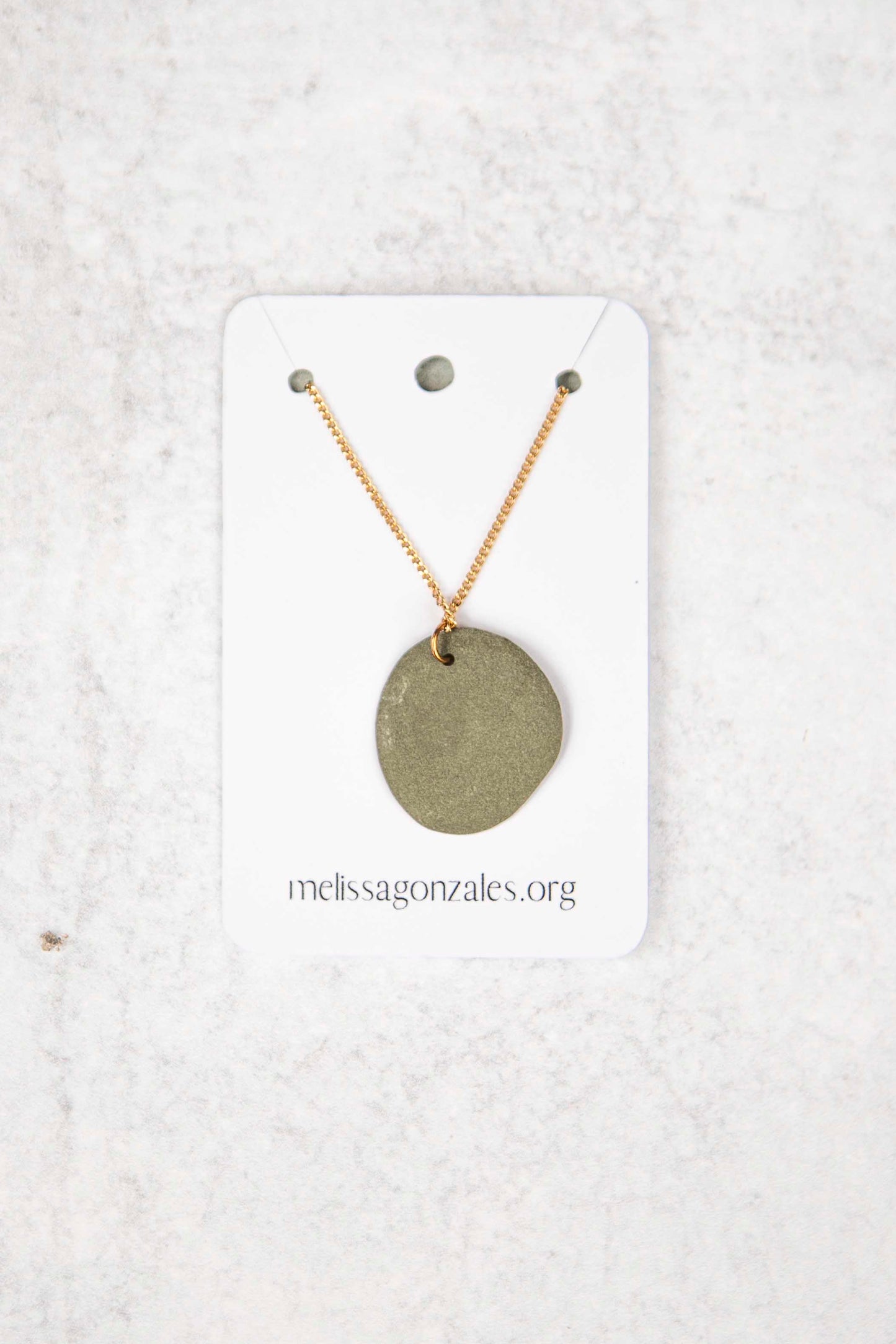 Necklace with a dark gray clay round pendant on a white card with 'melissagonzales.org' text, against a textured white background.